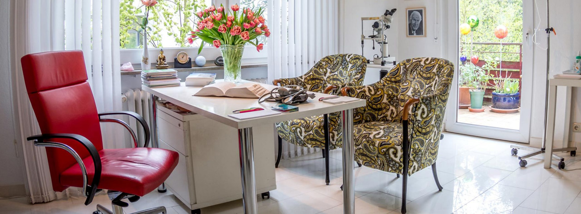 Beratungszimmer der Naturheilpraxis mit weißem Schreibtisch, rotem Sessel, gemusterten Sesseln und Tulpenstrauß