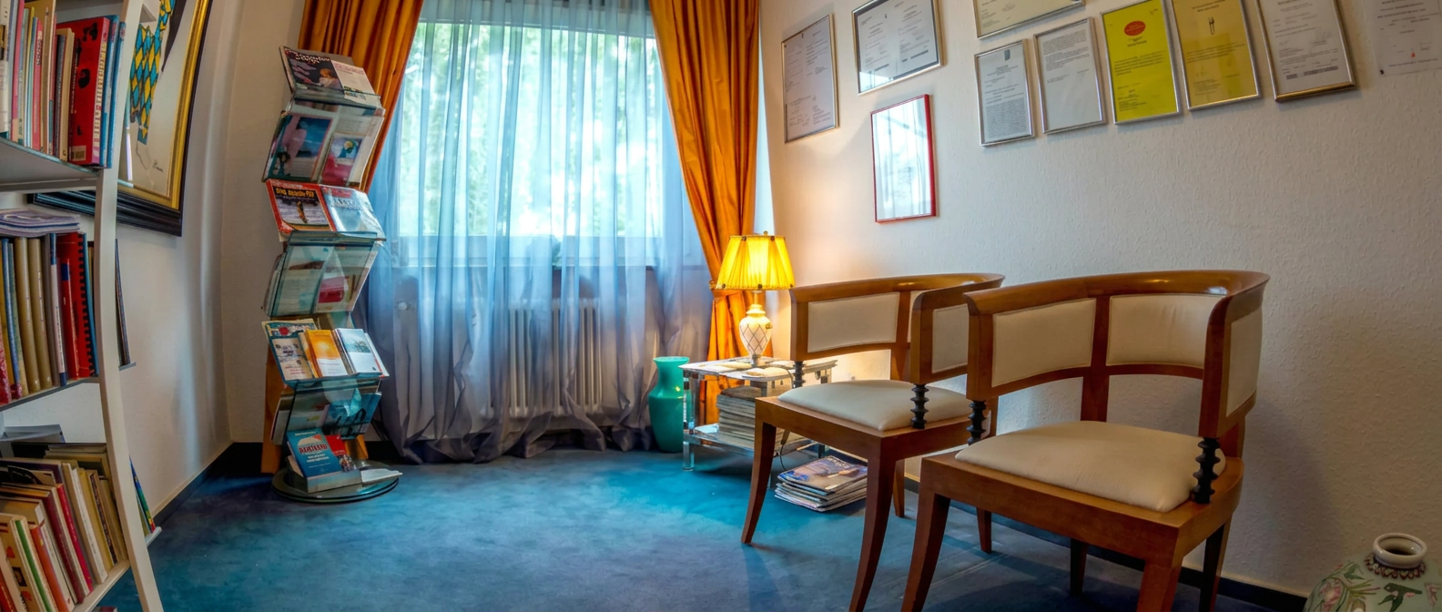 Wartezimmer der Praxis mit zwei Holzsesseln, Zeitschriftenständer, Bücherregal und Fenster mit orangefarbenen Vorhängen